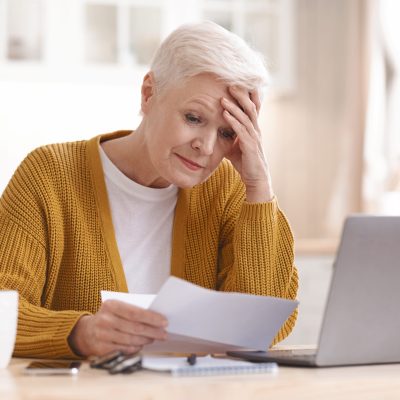 Upset senior lady holdign her head while reading bills, having financial troubles during quarantine. Sad elderly woman sitting in kitchen in front of laptop and holding documents, copy space